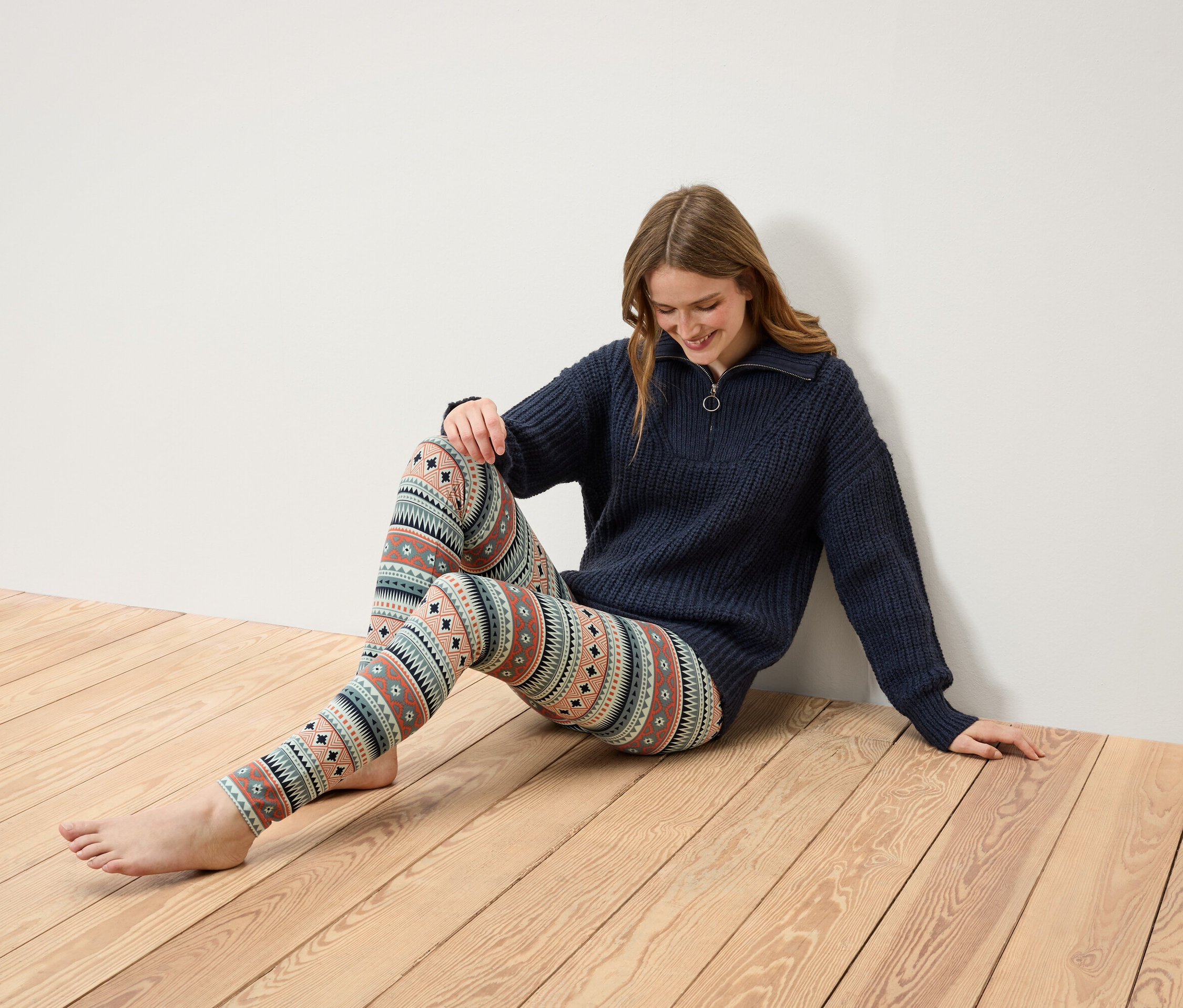Kobieta siedzi na drewnianej podłodze, ubrana w granatowy sweter na zamek i wzorzyste legginsy.