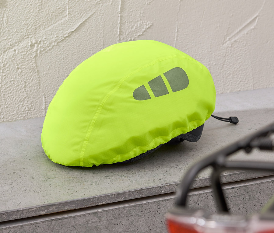 Żółty pokrowiec na kask z elementami odblaskowymi leży na szarej półce, w tle widać czerwone światło roweru.