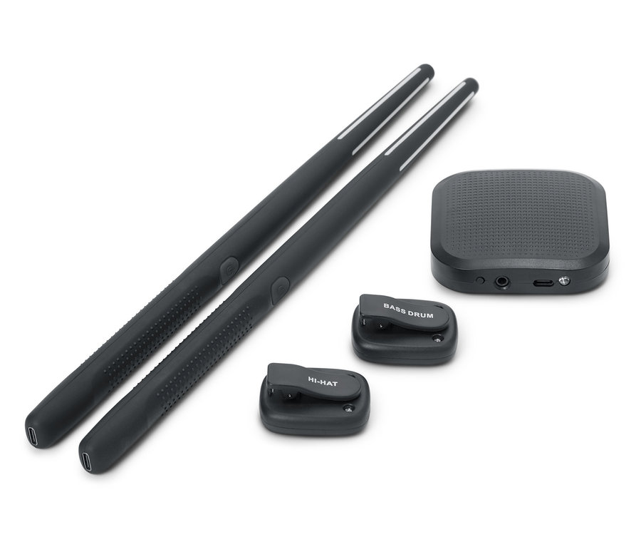 Dwie czarne pałeczki perkusyjne leżą obok siebie. Dostępny jest również czarny soundbox i dwa małe czarne pady perkusyjne, jeden z napisem BASS DRUM, a drugi z napisem HI-HAT.