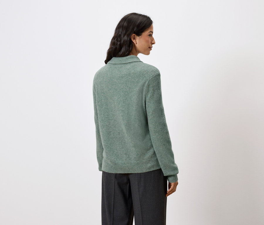 Widok z tyłu kobiety z ciemnymi włosami, ubranej w zielony sweter i ciemne spodnie.