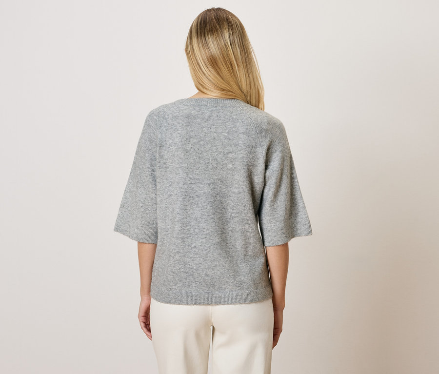 Z tyłu blondynka w szarym swetrze z rękawami trzy czwarte i białych spodniach.