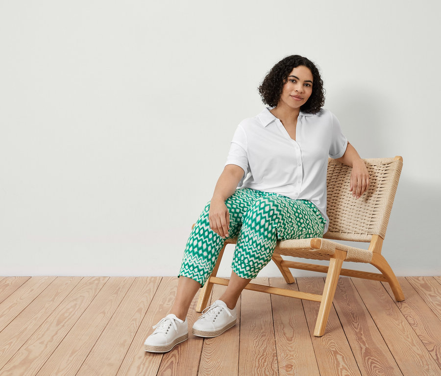 Kobieta siedzi na krześle i ma na sobie wzorzyste spodnie dżersejowe o długości 3/4 i białą bluzkę zapinaną na guziki.