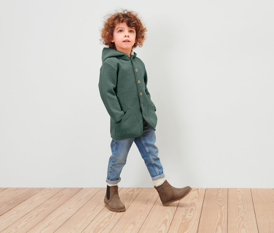 Chłopiec z kręconymi włosami idzie po drewnianej podłodze, ubrany w zielony wełniany płaszcz z kapturem, dżinsy i brązowe buty.