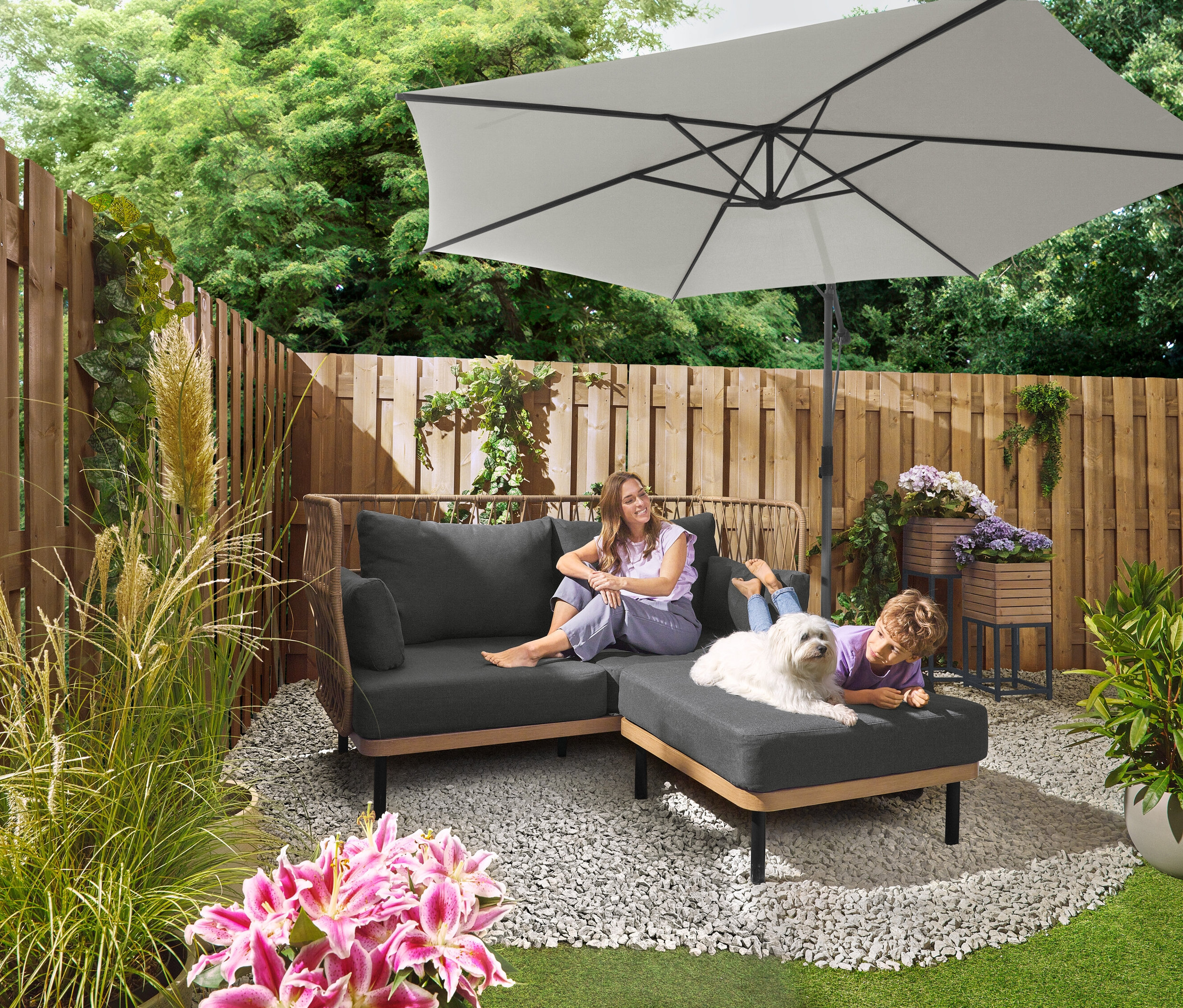 Kobieta siedzi na szarej sofie pod parasolem Leco Ampelschirm Ø 300 cm z podstawą i pokrowcem. Dziecko leży z psem na otomanie. W tle widoczny jest zestaw płyt Leco do stojaka krzyżowego.