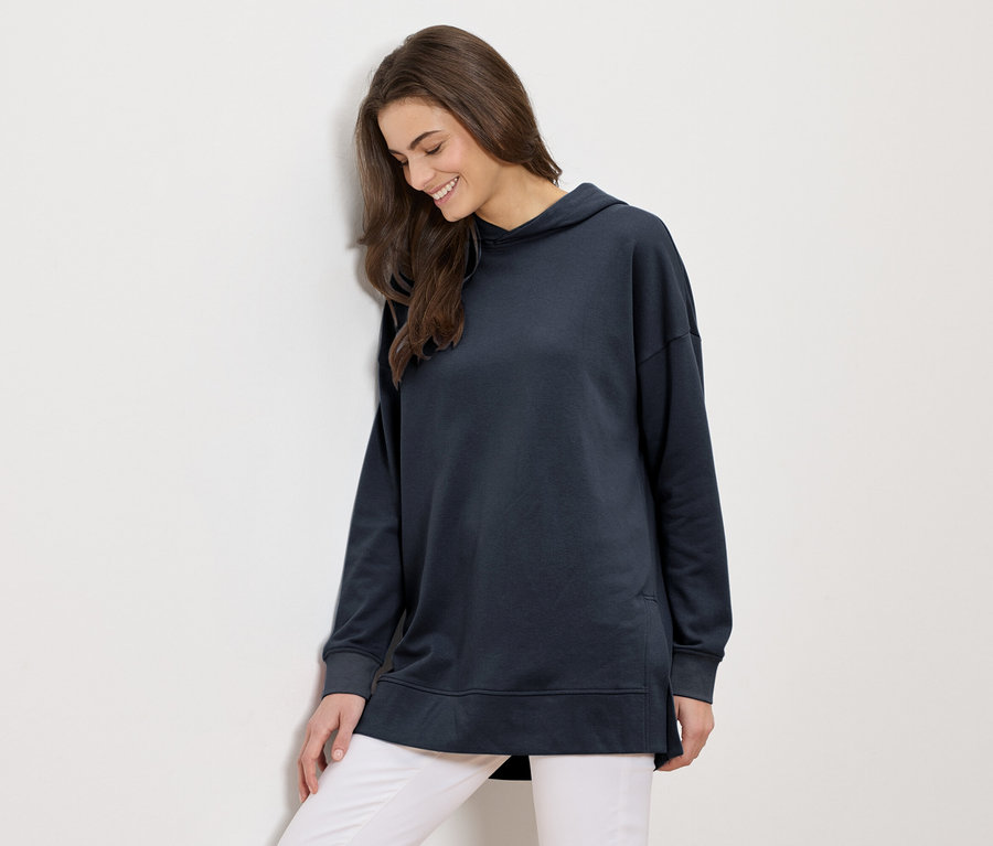 Kobieta z długimi brązowymi włosami w długiej granatowej bluzie z kapturem i białych spodniach uśmiecha się i opiera o białą ścianę.