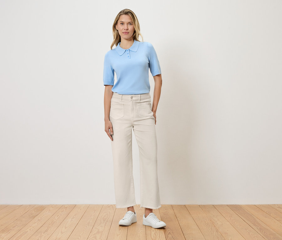 Kobieta stoi na tle ściany, ubrana w jasnoniebieską koszulkę polo, białe spodnie i białe buty.