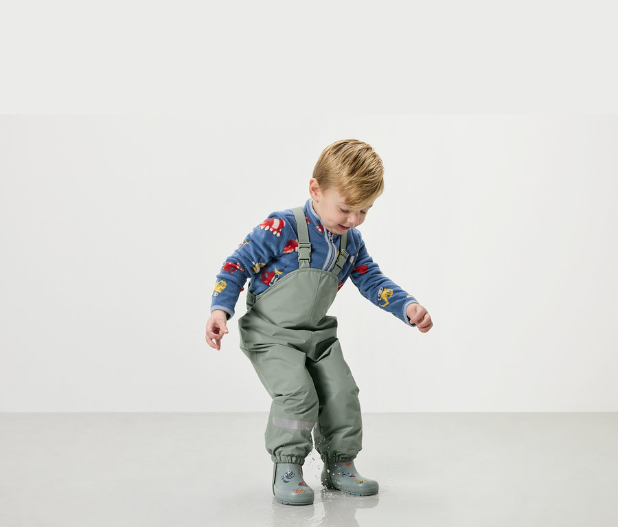 Chłopiec bawi się w wodzie w zielonym ubraniu przeciwdeszczowym i kaloszach.