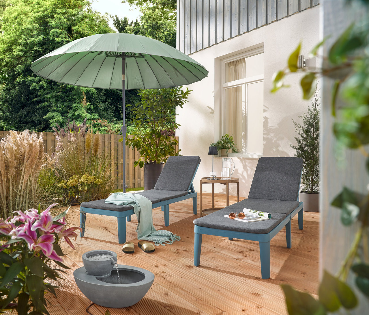 Na tarasie stoją dwa leżaki pod jasnoszarym parasolem Leco Shanghai i obok solarnej fontanny ogrodowej.