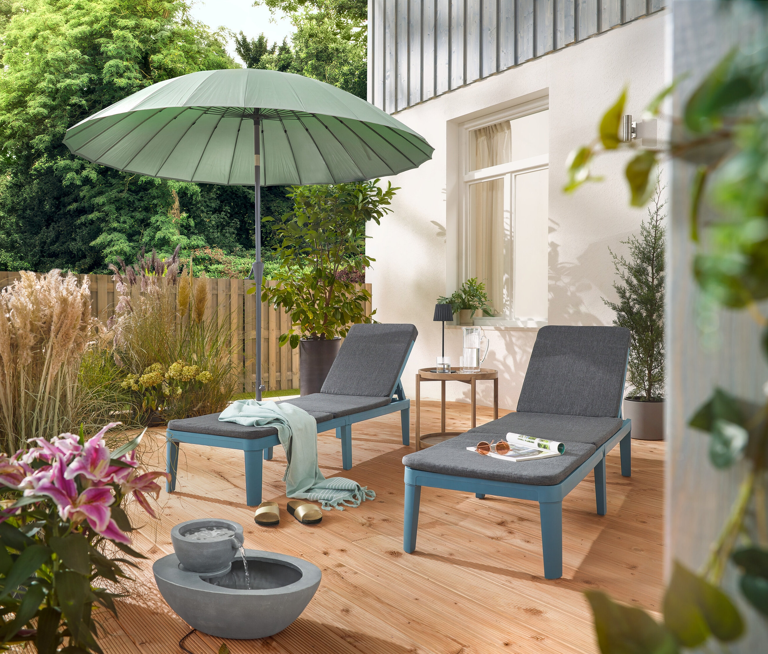 Na tarasie stoją dwa leżaki pod jasnoszarym parasolem Leco Shanghai i obok solarnej fontanny ogrodowej.