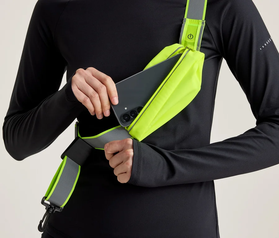 Osoba w czarnym longsleeve'ie wkłada czarny smartfon do neonowo żółtej torby na ramię z odblaskowym elementem.