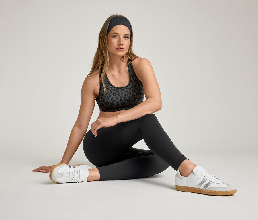 Kobieta siedzi w staniku sportowym, opasce i legginsach sportowych „ActiveSoft”. Nosi białe tenisówki.