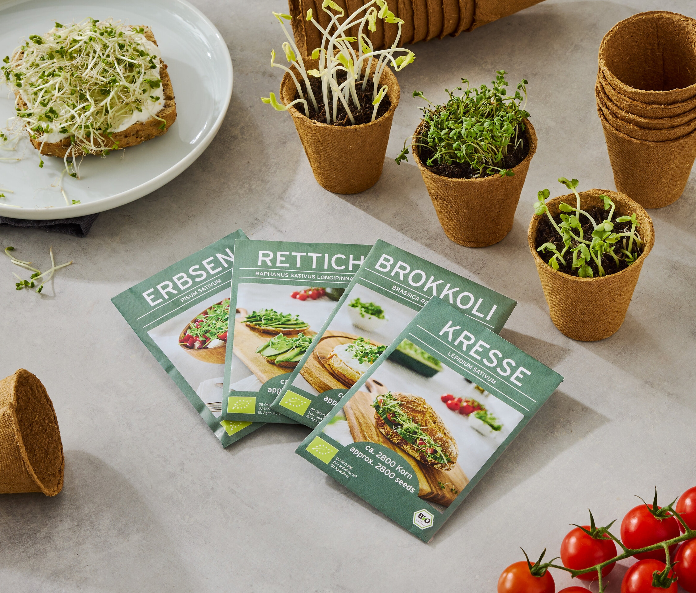 Talerz z chlebem posypanym kiełkami, wraz z czterema pudełkami nasion (grochu, rzodkiewki, brokułów, rzeżuchy), małymi doniczkami z rosnącymi kiełkami i gałązką pomidorów.