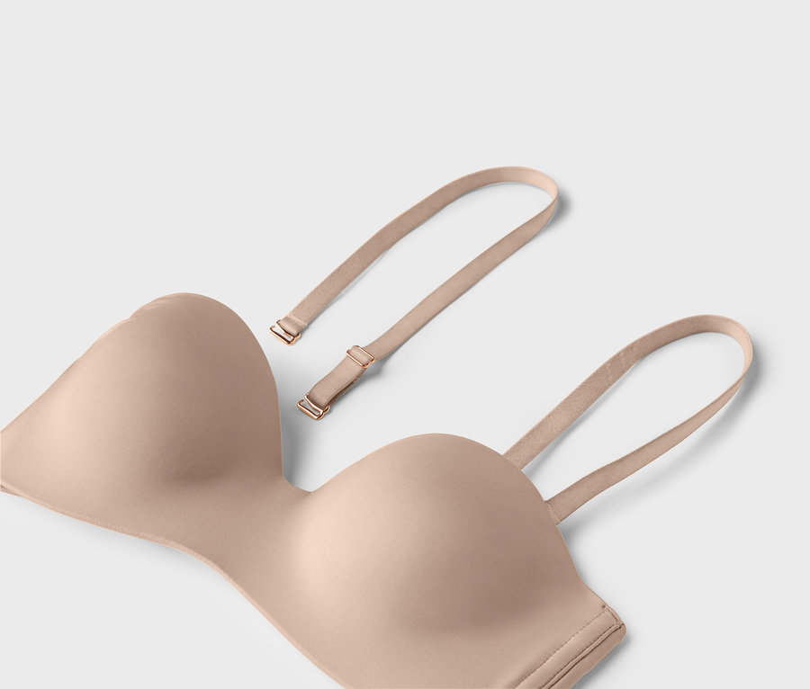 Beżowy biustonosz bez ramiączek z odpinanymi ramiączkami leży na jasnym tle.