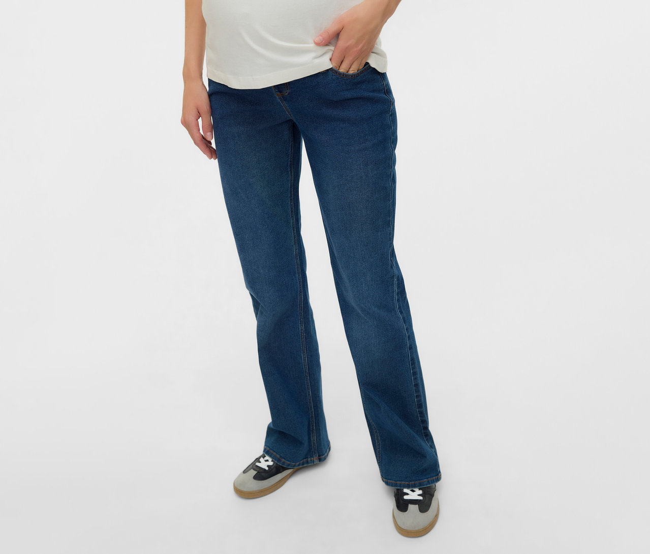 Kobieta w ciąży w rozszerzanych jeansach MAMALICIOUS „MLEDINA” i białej koszulce, z ręką w kieszeni.