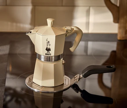 Kremowa kawiarka do espresso Bialetti Moka Express 190 ml stoi na adapterze do płyt indukcyjnych Bialetti 13 cm obok szklanki espresso.