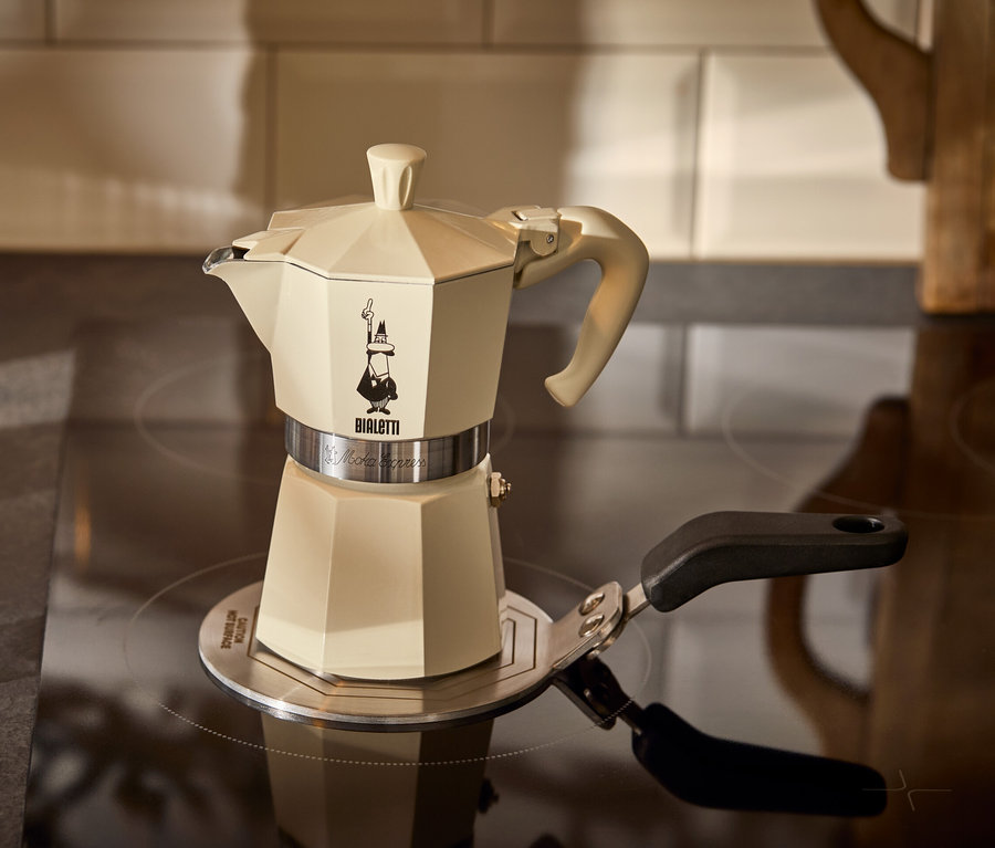 Kremowa kawiarka do espresso Bialetti Moka Express 190 ml stoi na adapterze do płyt indukcyjnych Bialetti 13 cm obok szklanki espresso.