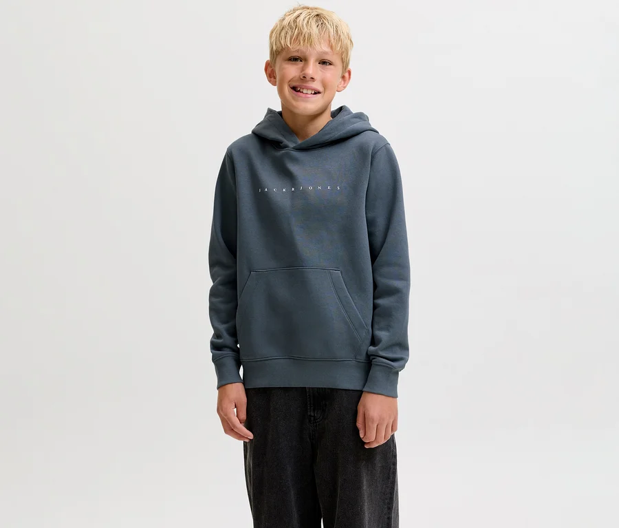 Uśmiechnięty chłopiec w ciemnoszarej bluzie z kapturem Jack&Jones Star z kieszenią kangurką i ciemnymi spodniami.