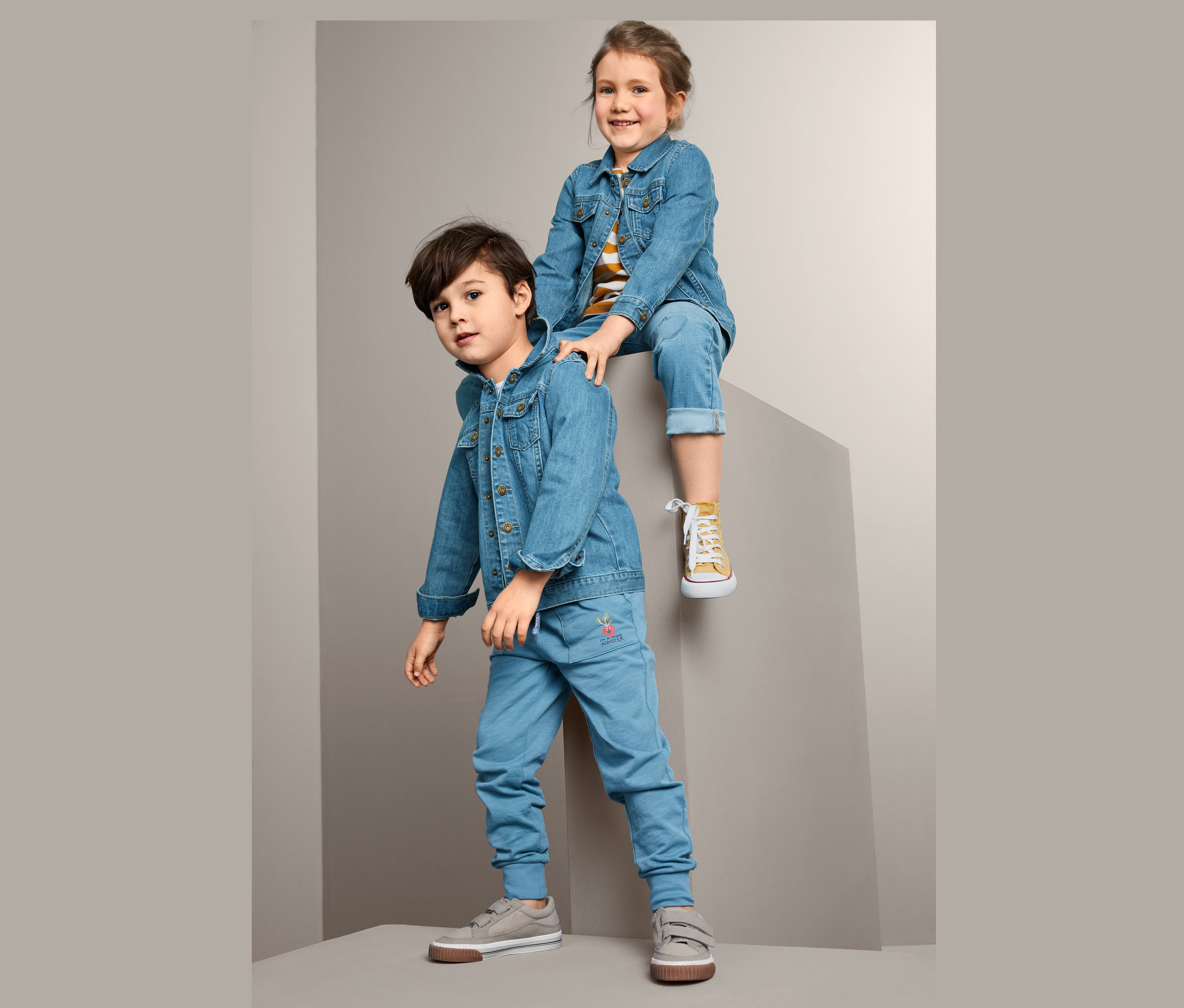 Chłopiec stoi, a dziewczynka siedzi na szarym bloku, oboje w kurtkach dżinsowych.