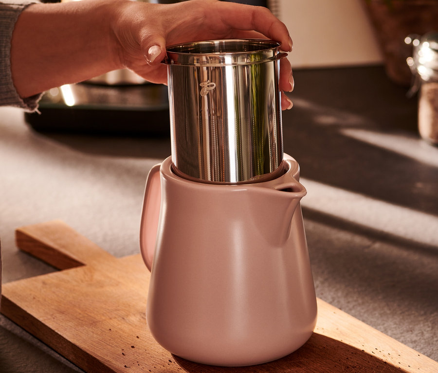 Ręka trzyma filtr ze stali nierdzewnej nad ceramicznym zaparzaczem do kawy w kolorze różowym.