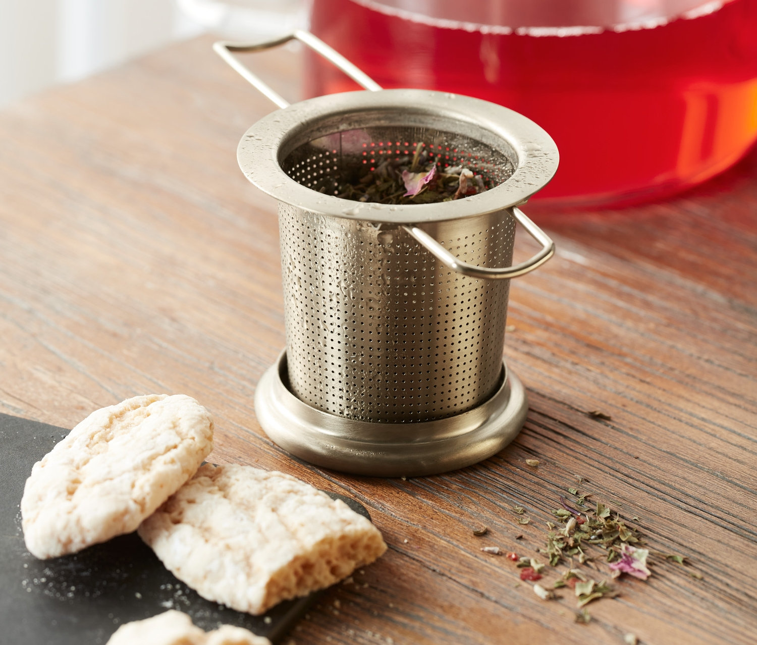 Zaparzacz do herbaty ze stali nierdzewnej z ziołami stoi na drewnianym stole obok ciasteczek i dzbanka z czerwonym napojem.