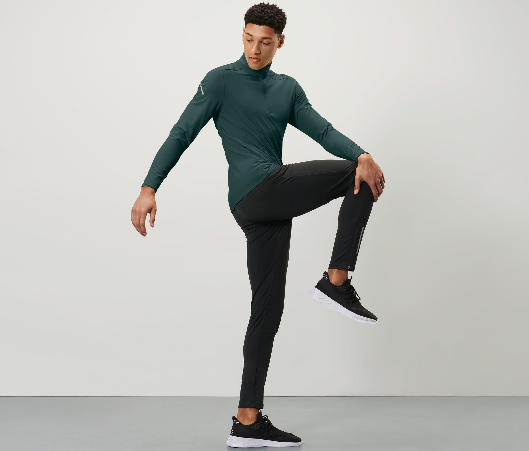 Mężczyzna w zielonej koszulce sportowej i czarnych legginsach do biegania chroniących przed wiatrem trzyma jedno kolano ręką przed klatką piersiową.