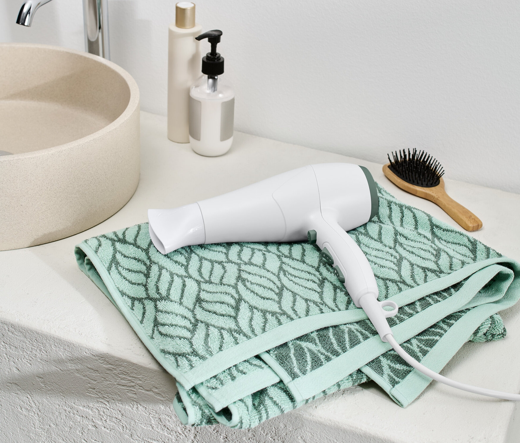 Biała suszarka do włosów z jonizacją leży na zielonym ręczniku w łazience.