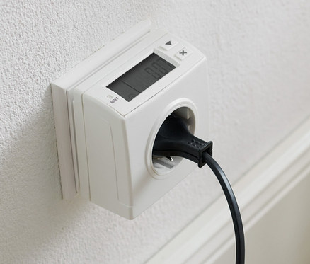 Na białej ścianie znajduje się gniazdko z licznikiem zużycia energii i podłączonym czarnym sznurem.