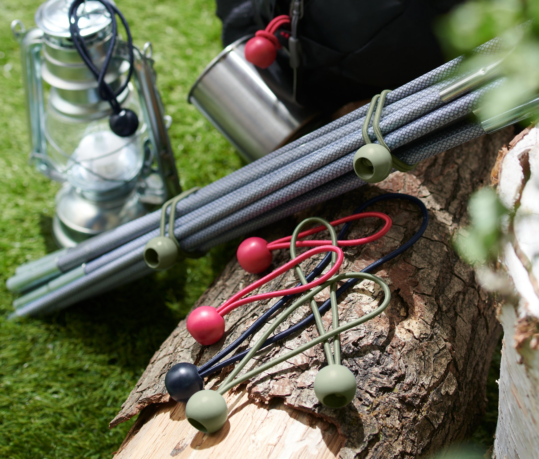 Na kłodzie leży 12 gumek napinających z kulkami, obok nich lampion, metalowy kubek, tyczki i plecak.