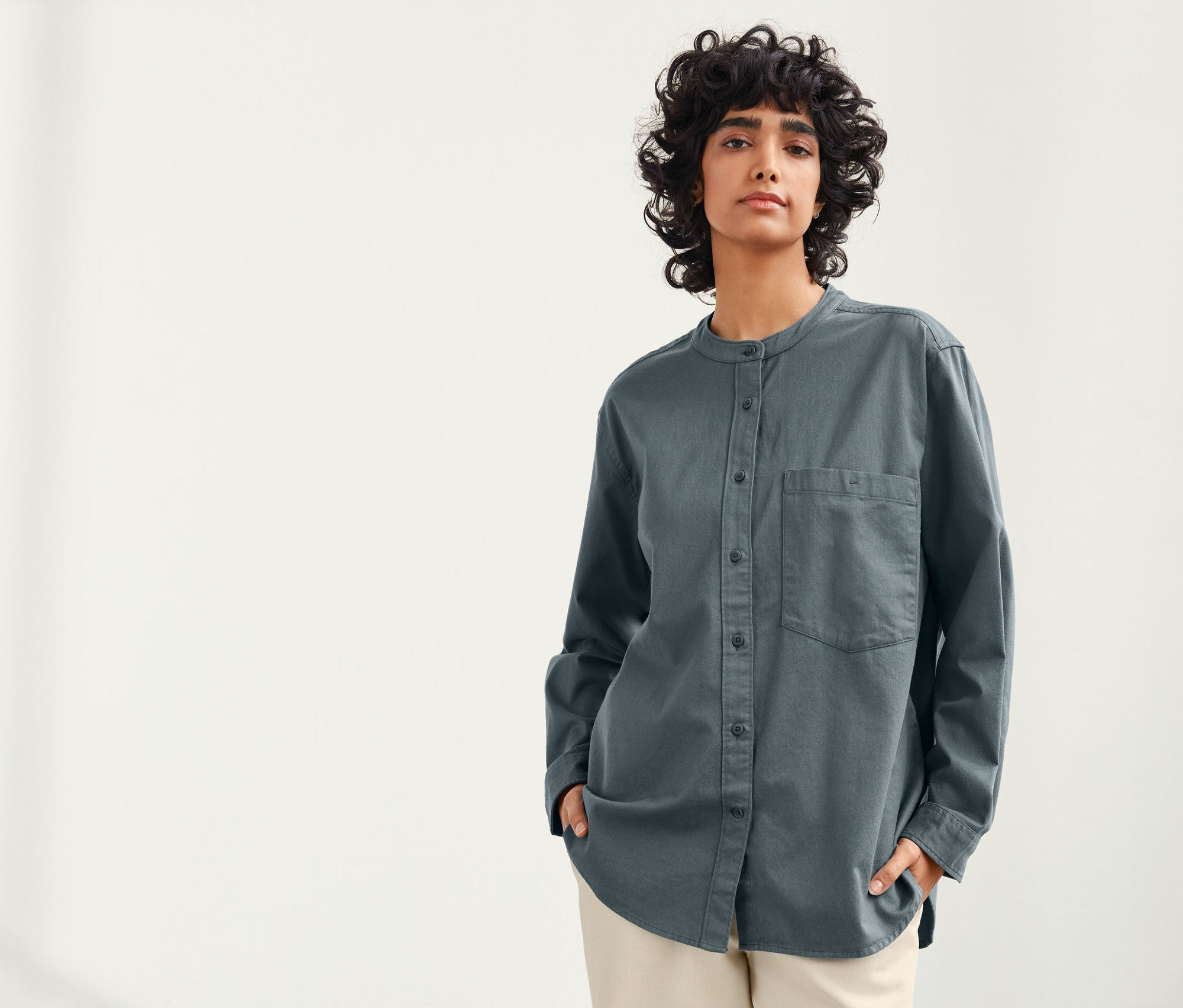 Kobieta o ciemnych kręconych włosach pozuje na jasnym tle, ubrana w niebiesko-szarą lnianą bluzkę NAH/STUDIO.