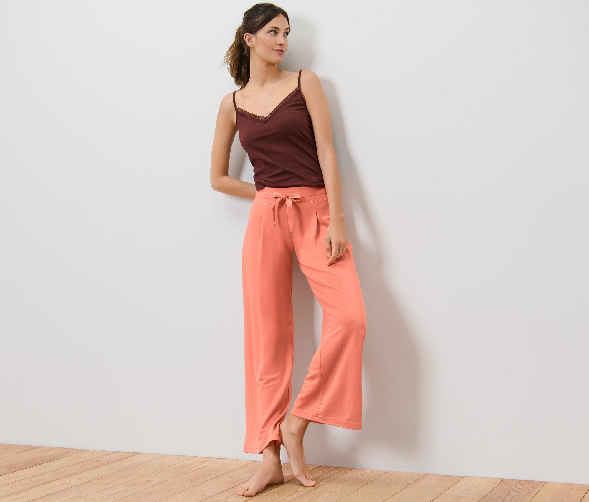 Kobieta opiera się o ścianę, ubrana w Spodnie rekreacyjne i brązowy top.