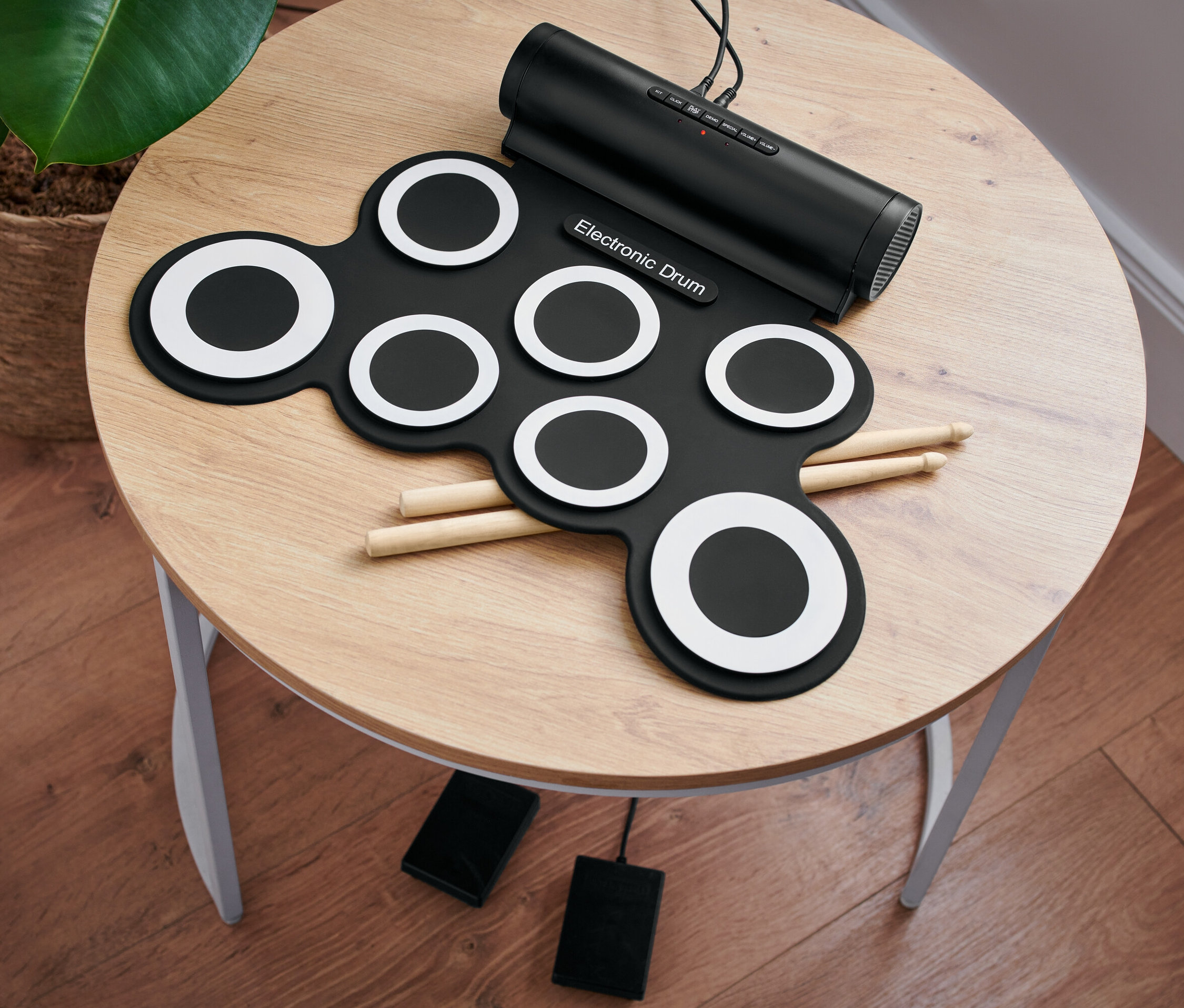 Na okrągłym stole leży elektroniczny zestaw perkusyjny z pałeczkami i pedałami.
