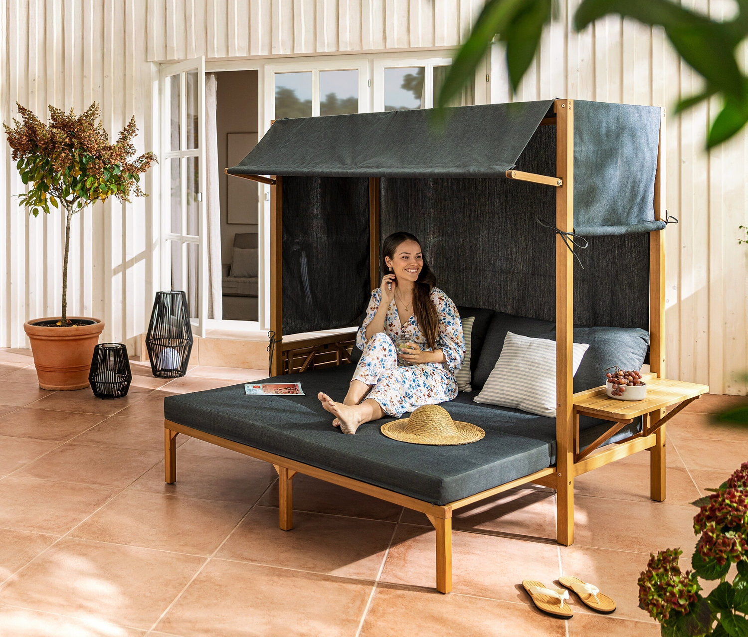 Kobieta siedzi w koszu plażowym „Tinus” z wysuwanym podnóżkiem. W tle widać stół rozkładany „Elin” i fotel do jadalni z plecionką tekstylną.