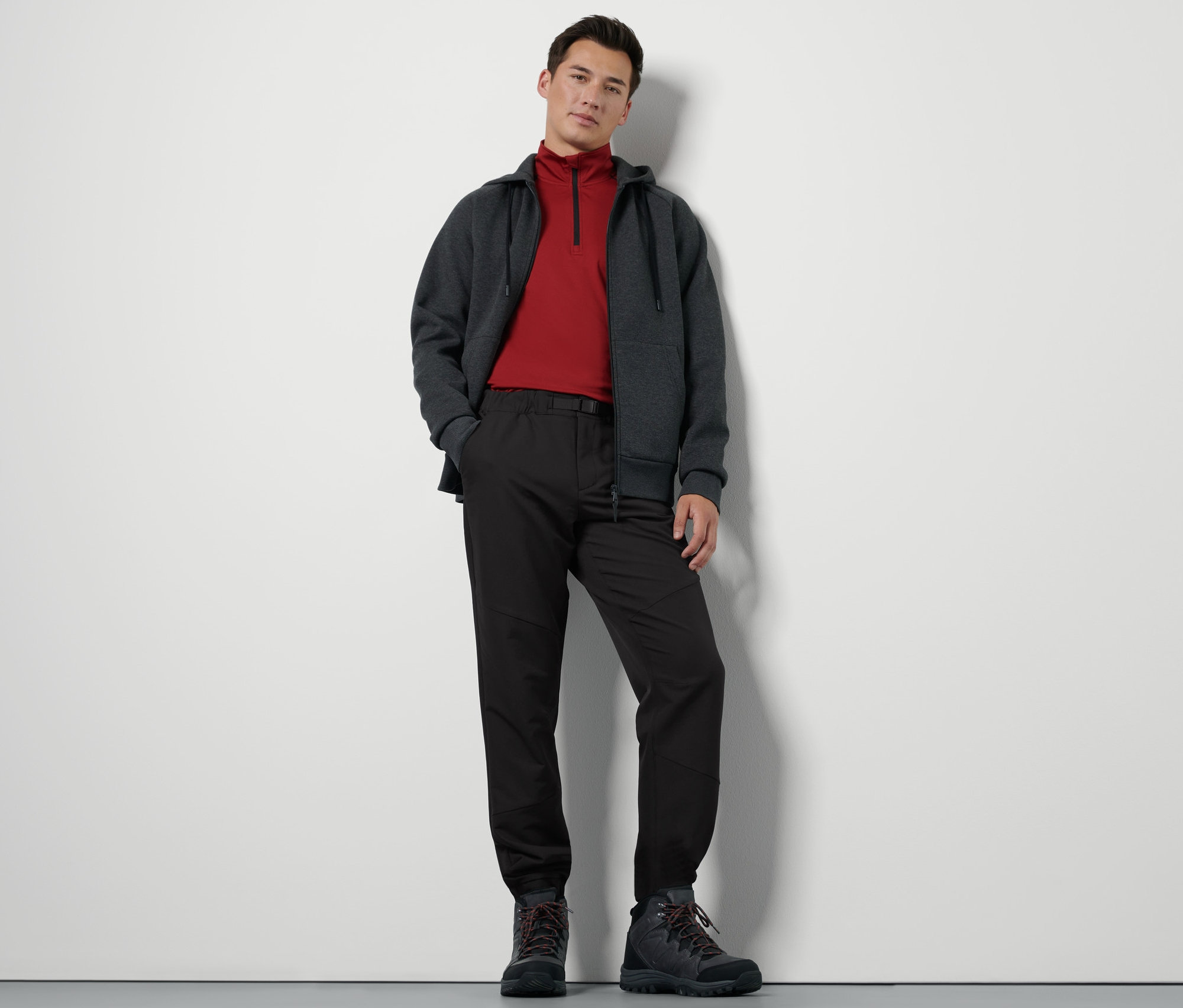 Mężczyzna stoi i ma na sobie spodnie termiczne z paskiem, bluzę z zamkiem błyskawicznym i buty turystyczne.