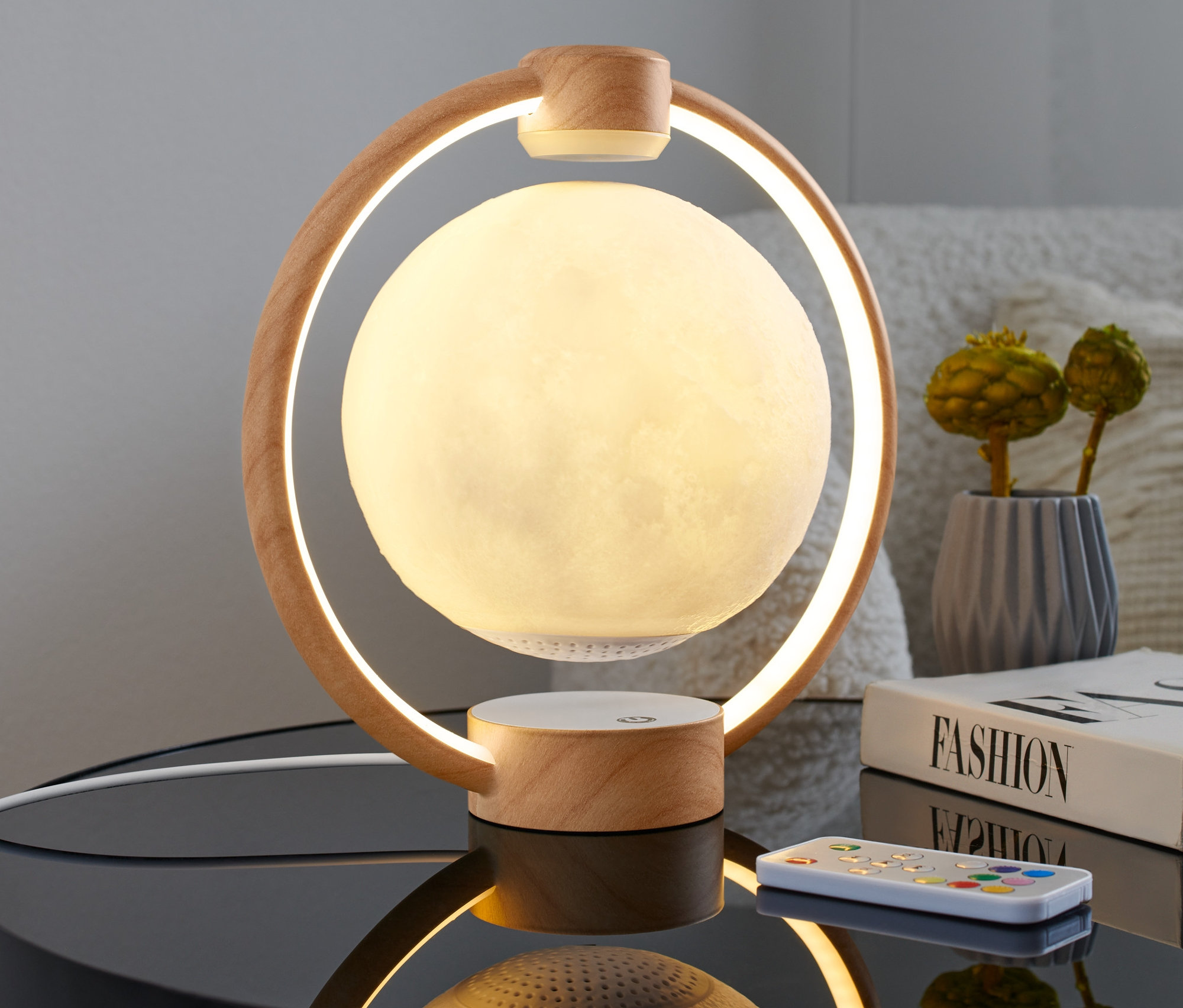 Głośnik LED Bluetooth® z unoszącym się w powietrzu księżycem stoi na stoliku nocnym obok książki i pilota.