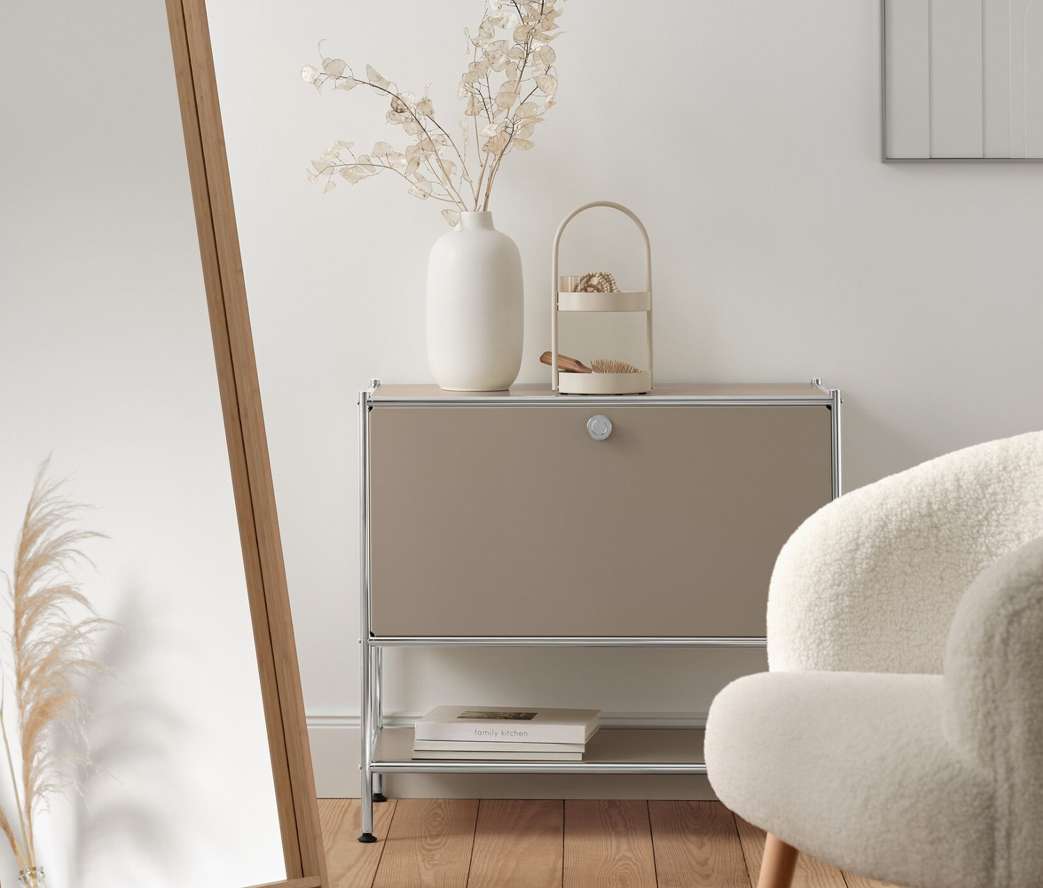 Wnętrze z beżową metalową szafką typu sideboard „CN3” z klapą, lustrem stojącym z bambusową ramą i fotelem z tkaniny bouclé z drewnianymi nogami.