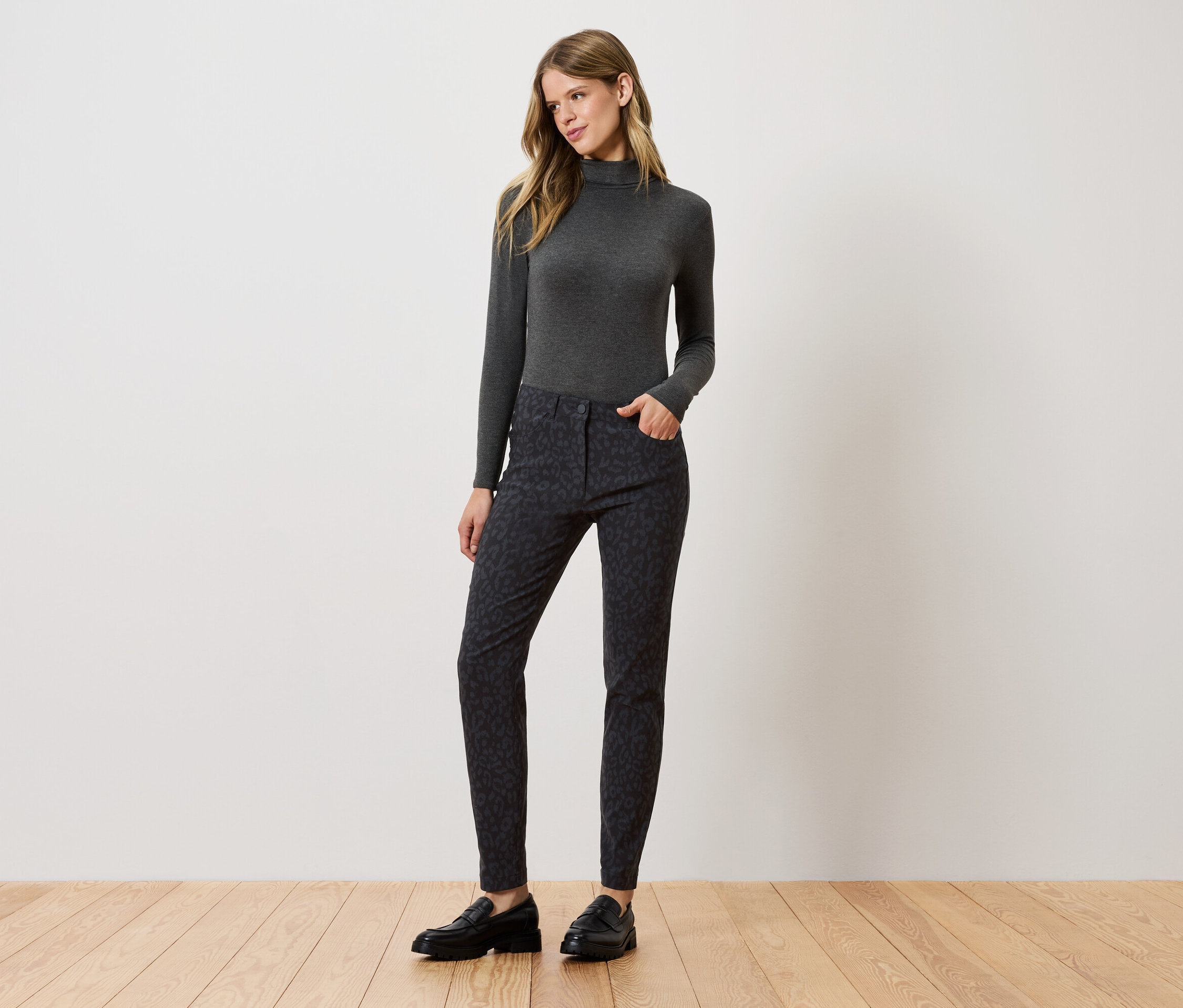 Kobieta stoi i pozuje w szarym golfie, wzorzystych spodniach i czarnych butach.
