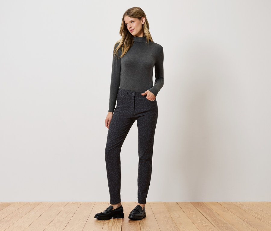 Kobieta stoi i pozuje w szarym golfie, wzorzystych spodniach i czarnych butach.