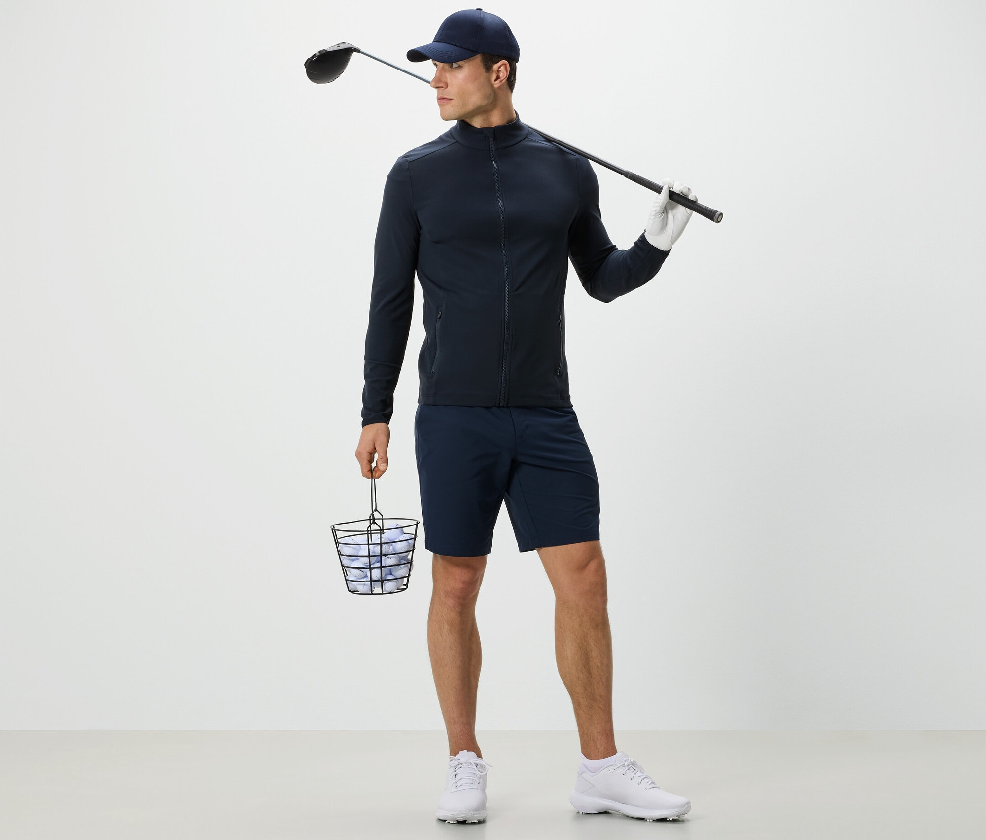 Mężczyzna w granatowej funkcyjnej bluzie trzyma kosz z piłkami golfowymi i kij golfowy.