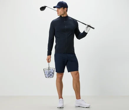 Mężczyzna w granatowej funkcyjnej bluzie trzyma kosz z piłkami golfowymi i kij golfowy.