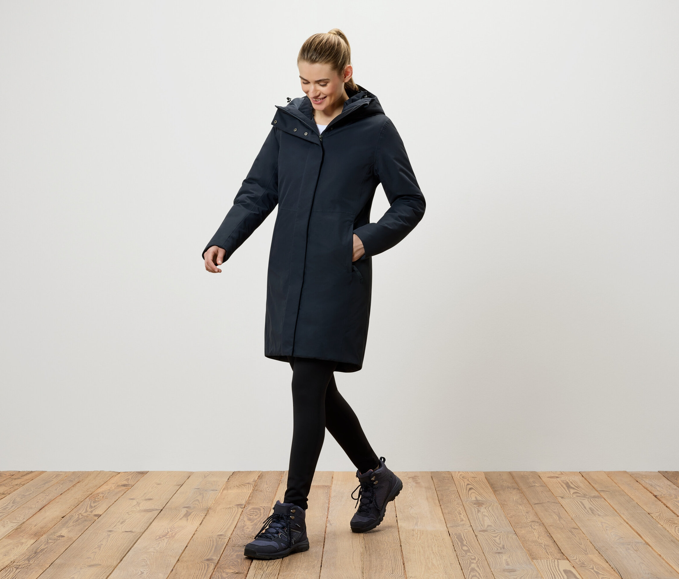 Kobieta idzie po drewnianej podłodze, ubrana w granatowy płaszcz z kapturem, czarne legginsy i granatowe buty.