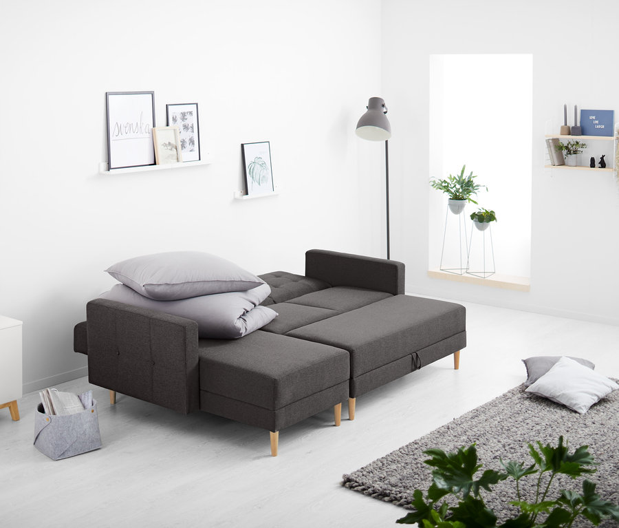 W salonie stoi sofa narożna sprężynowa ze skrzynią, na której leżą poduszki i koc.