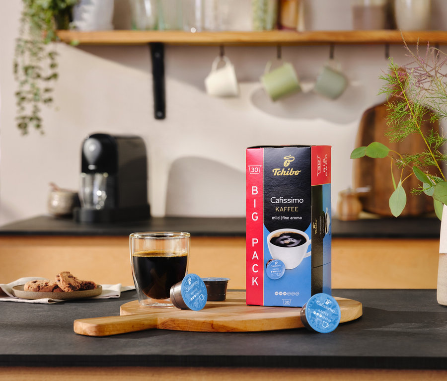 Na blacie stoi szklanka kawy obok opakowania Cafissimo pure+, black i dwóch kapsułek.