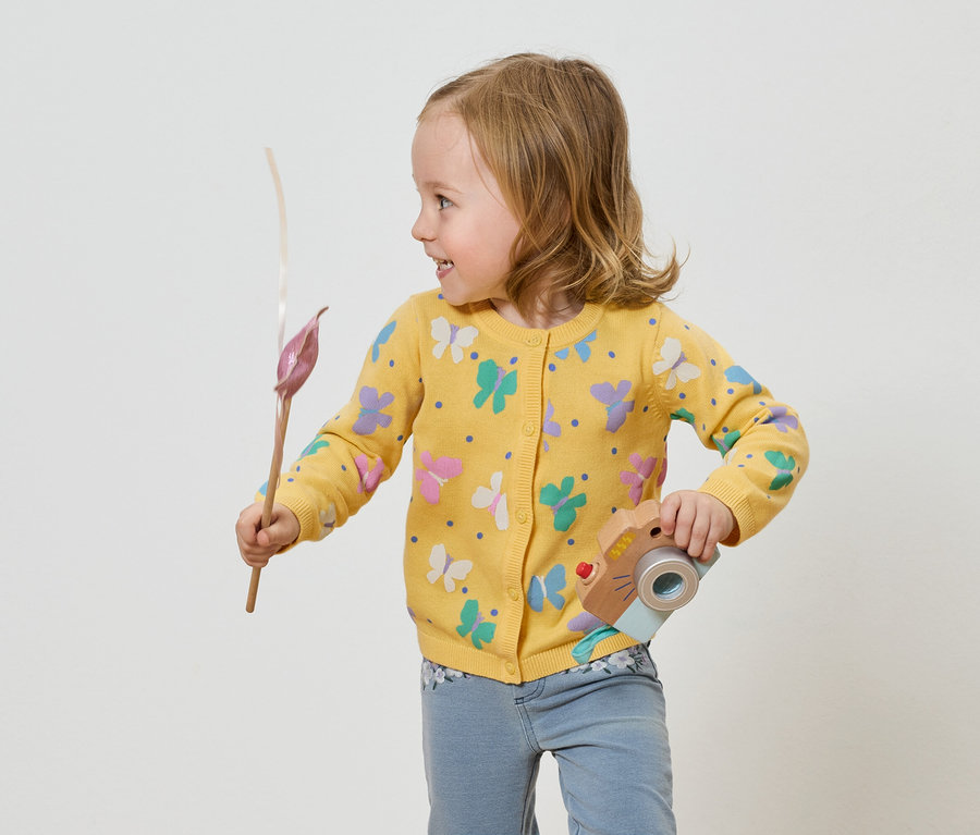 Dziewczynka w żółtym swetrze w motyle trzyma zabawkę-aparat i kwiatka.