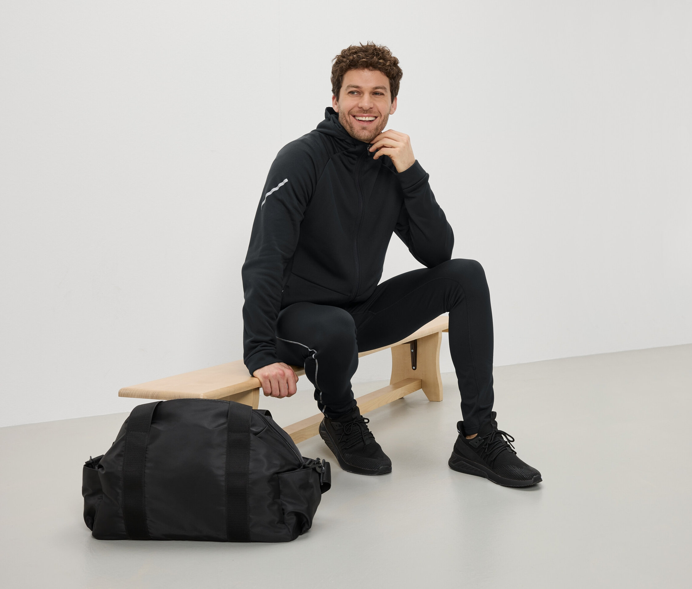 Mężczyzna siedzi na ławce w czarnym stroju sportowym obok czarnej torby sportowej.