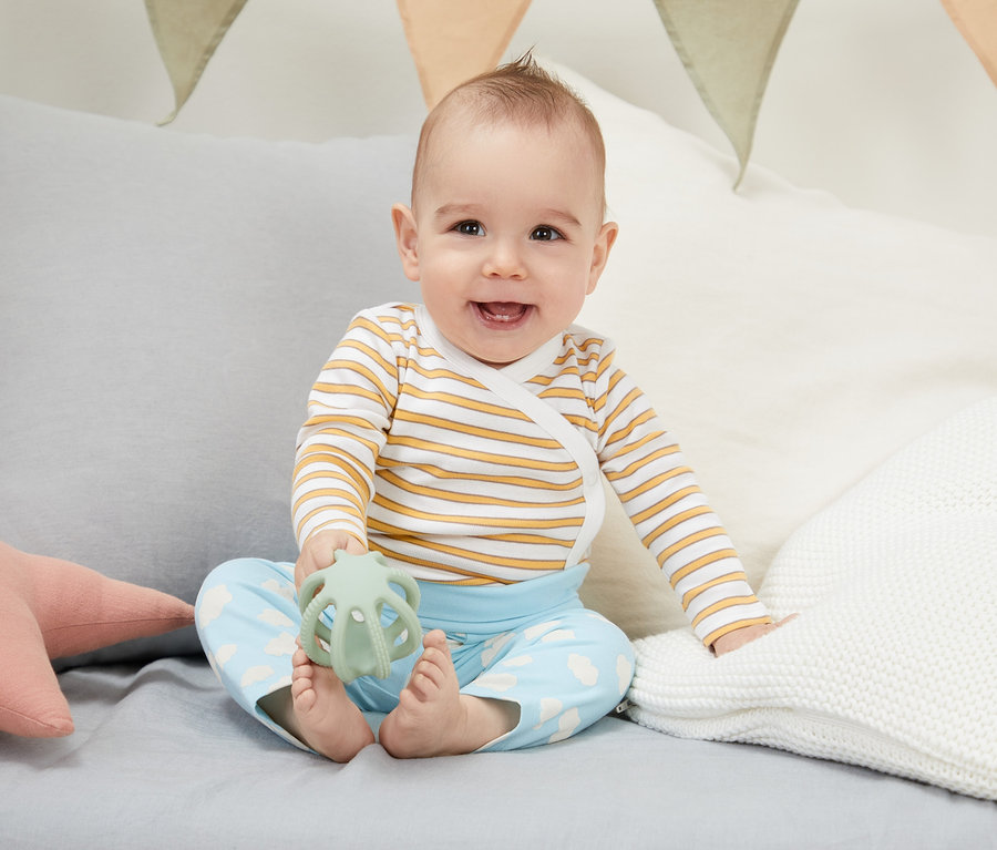 Uśmiechnięte dziecko siedzi na łóżku i trzyma zieloną zabawkę. Ma na sobie zestaw niemowlęcy z bawełny ekologicznej, 4-częściowy.