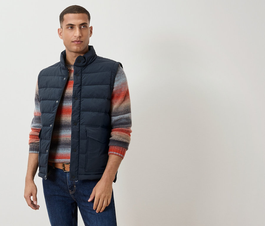 Mężczyzna ma na sobie granatową kamizelkę nałożoną na sweter w paski, brązowy pasek i dżinsy.