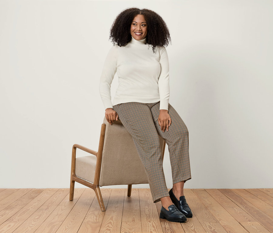 Kobieta siedzi na beżowym krześle, ubrana w biały golf, brązowe spodnie w pepitkę i czarne mokasyny.