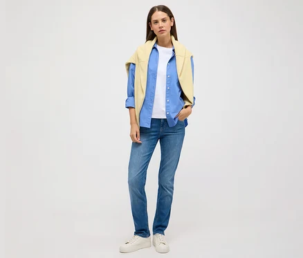 Kobieta stoi na jasnym tle i ma na sobie dżinsy Angels „Cici Push-Up”, Slim Fit, niebieską koszulę, biały T-shirt i żółty sweter.