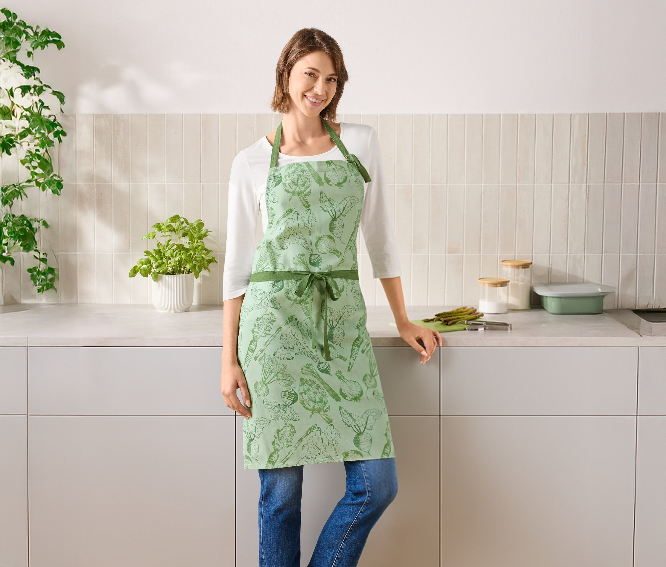 Kobieta stoi w kuchni, ubrana w zielony fartuch z motywem warzyw.