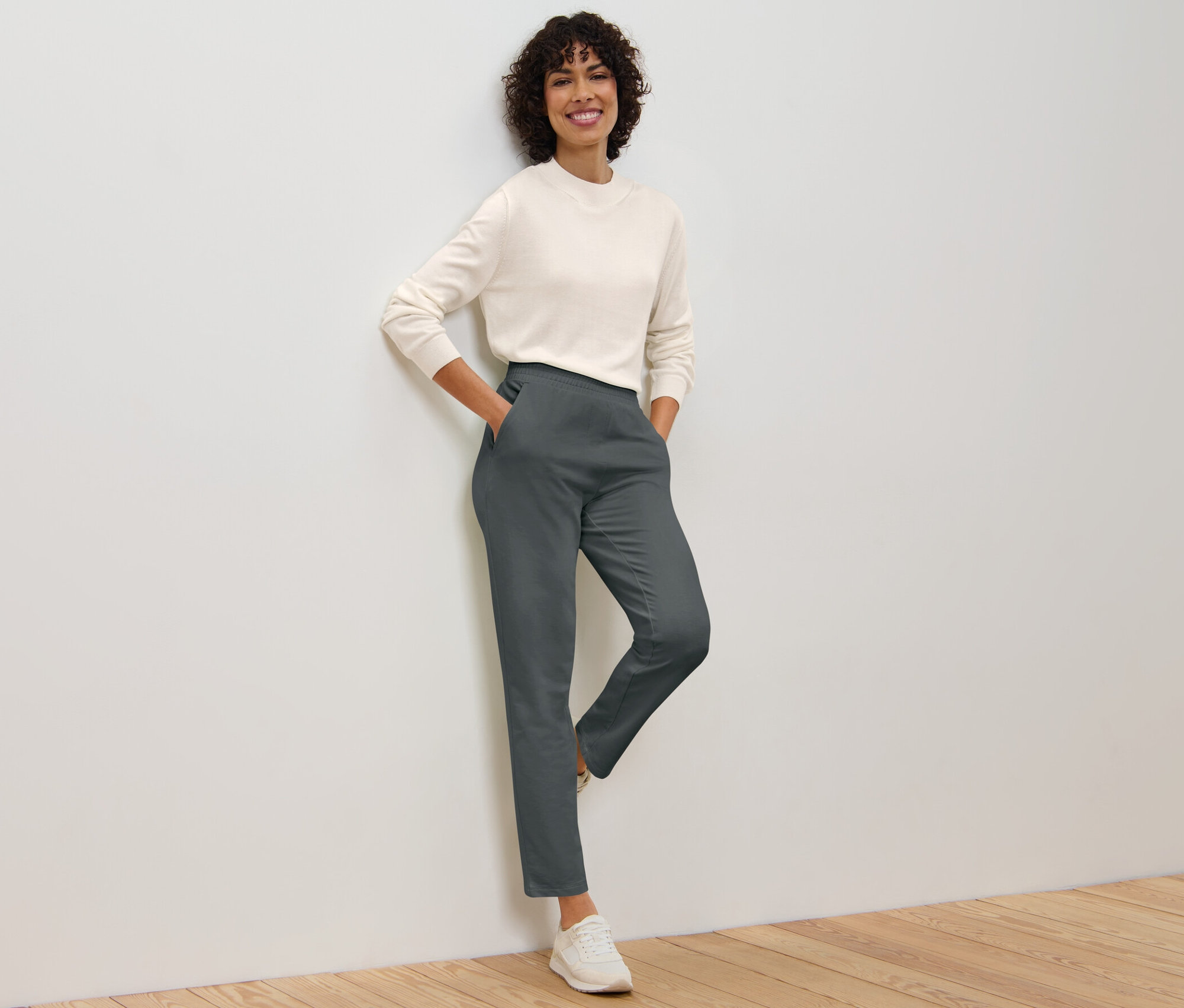 Kobieta oparta o ścianę pozuje w szarych spodniach joggery i swetrze.